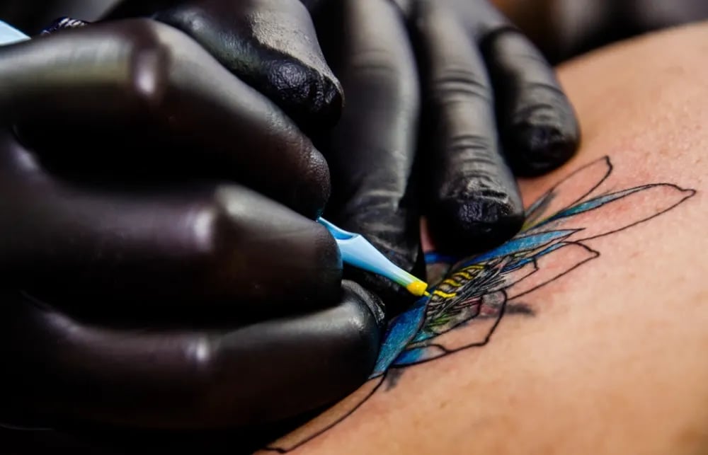 Gloved hand tattooing blue and yellow linework onto skin