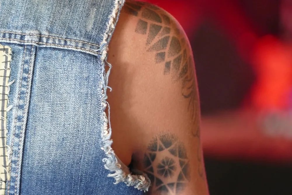 Dotwork geometric cap and mandala tattoos on a shoulder beside a denim vest