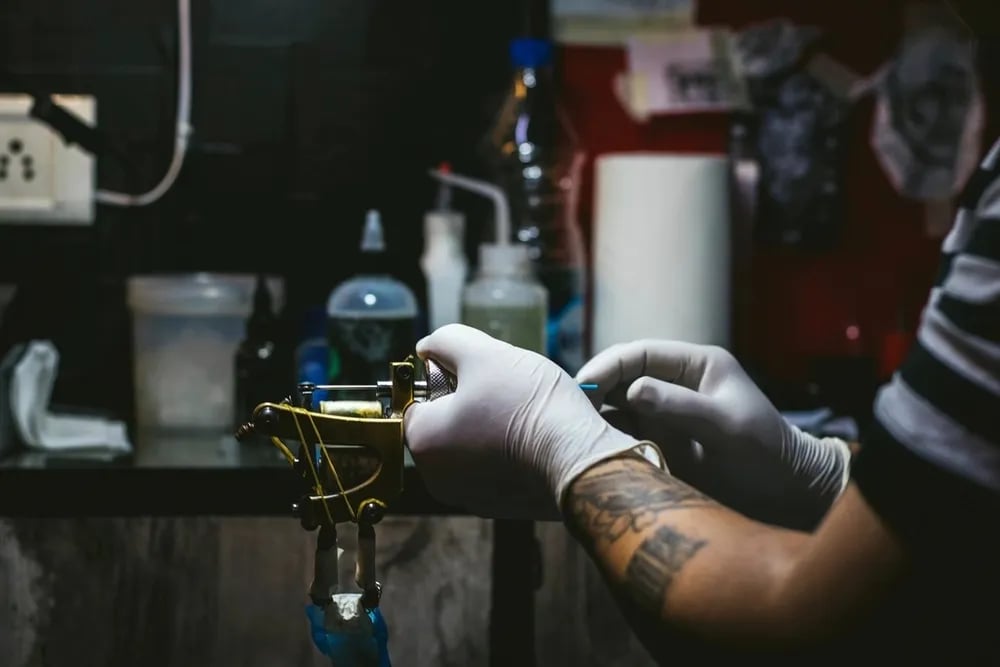 Tattooer in white gloves holding a coil machine at a prepared station