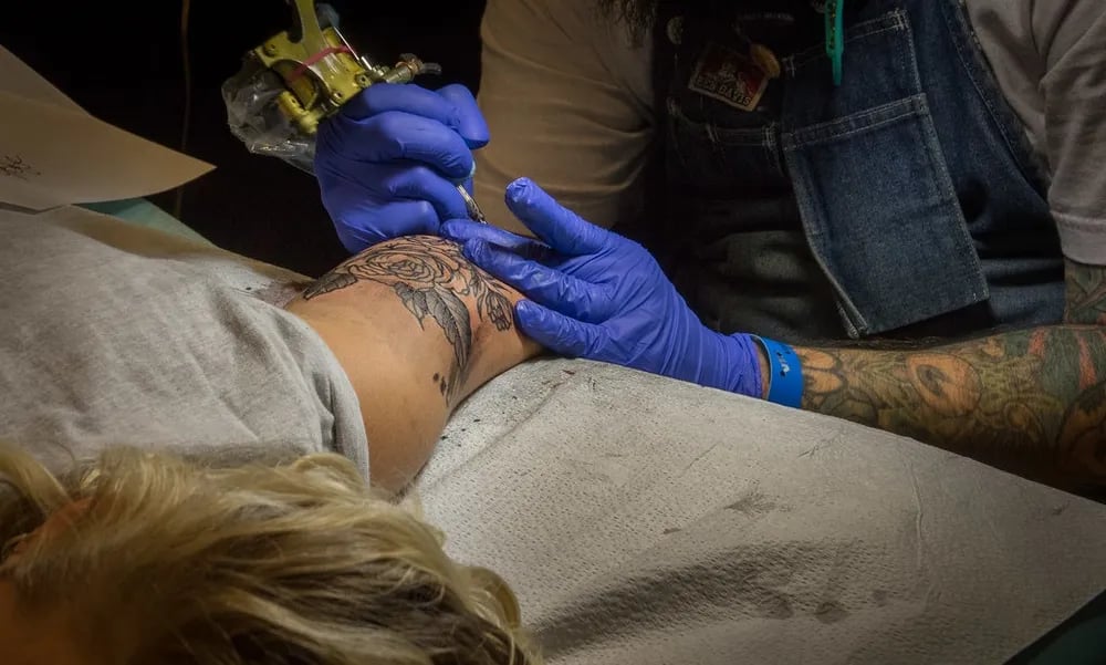 Client lying on a padded bench while a tattooer in blue gloves works on the back of her leg