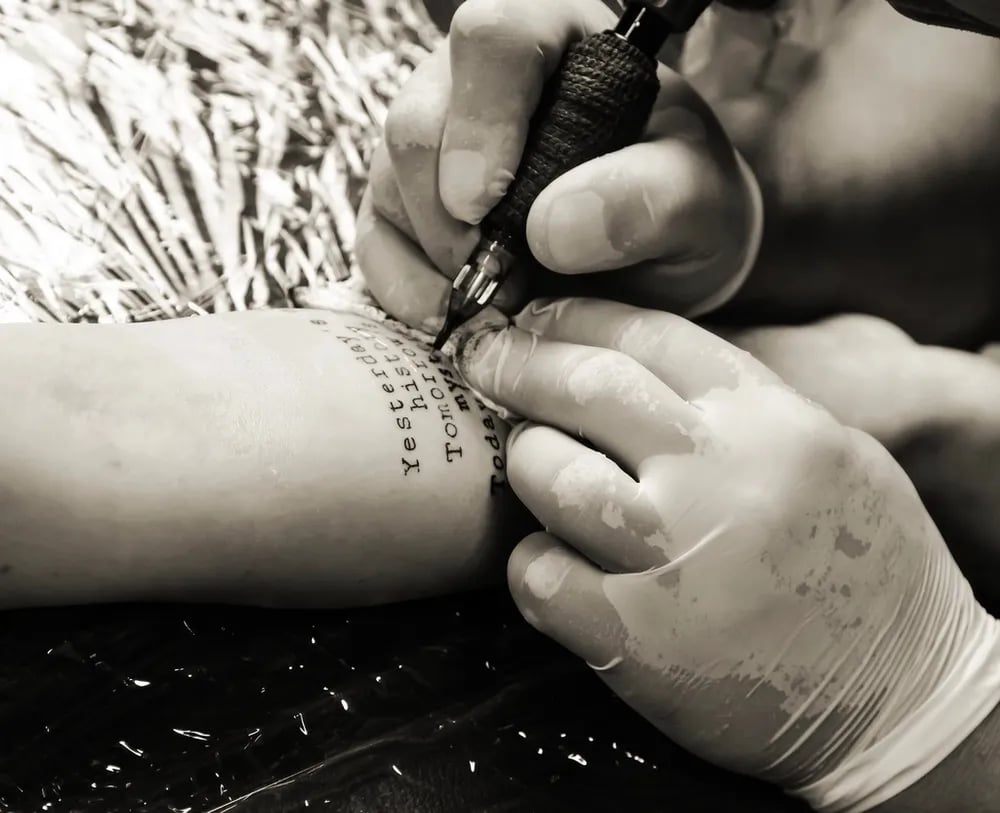 Black and white photo of gloved hands tattooing lines of lettering onto an arm
