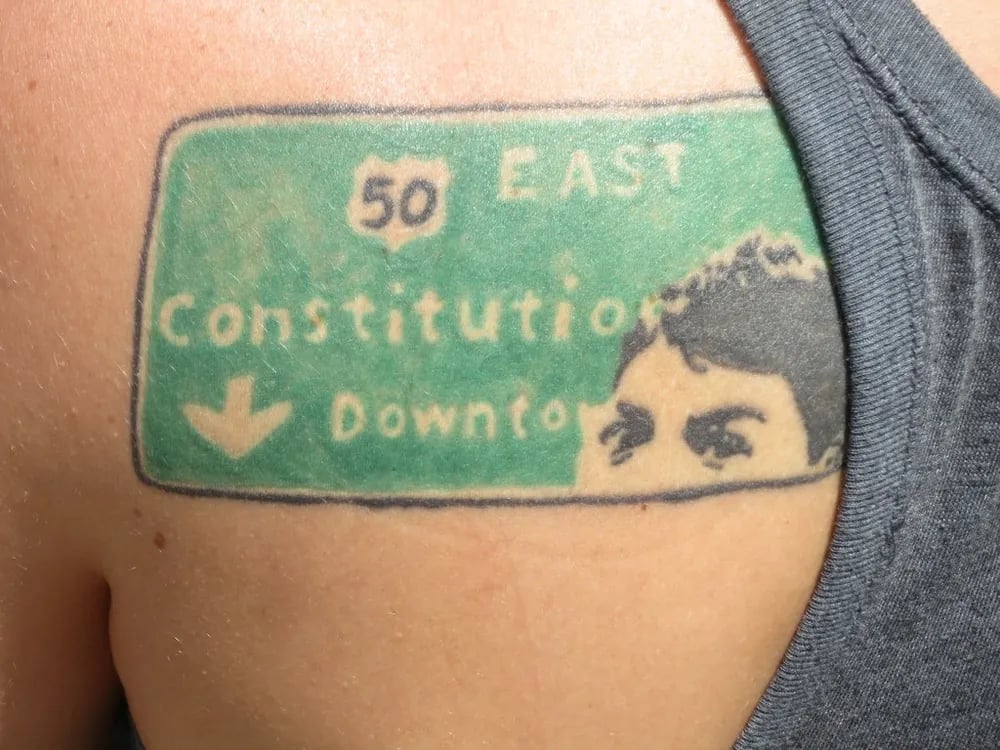 Green highway sign tattoo with a face peeking over its edge on a shoulder