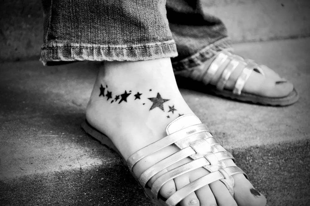 Black and white photo of a trail of small black stars tattooed across a foot in sandals