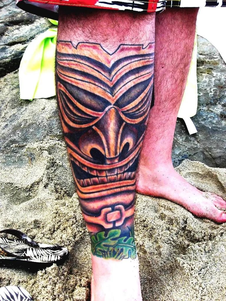Large tiki face tattoo in brown and red tones covering a calf, on a sandy beach