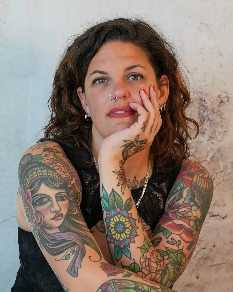 Woman resting her chin on her hand, both arms covered in colourful traditional patchwork tattoos