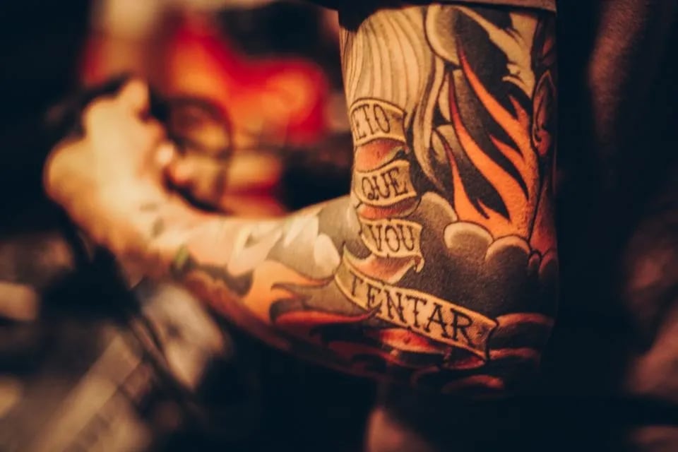 Colourful forearm tattoo of a lettered banner framed by flames, in warm light