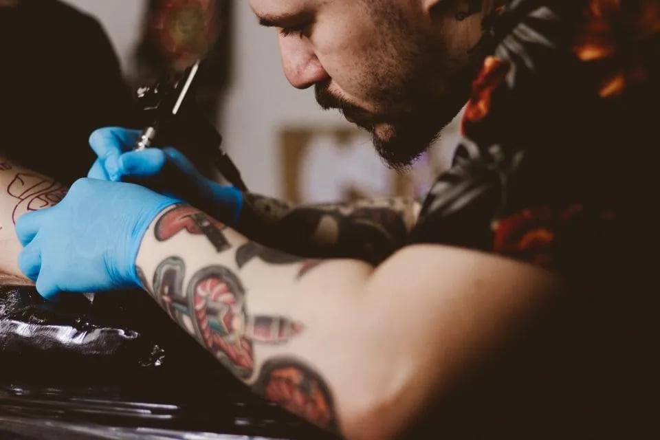 Bearded tattooer in blue gloves working on a colourful arm